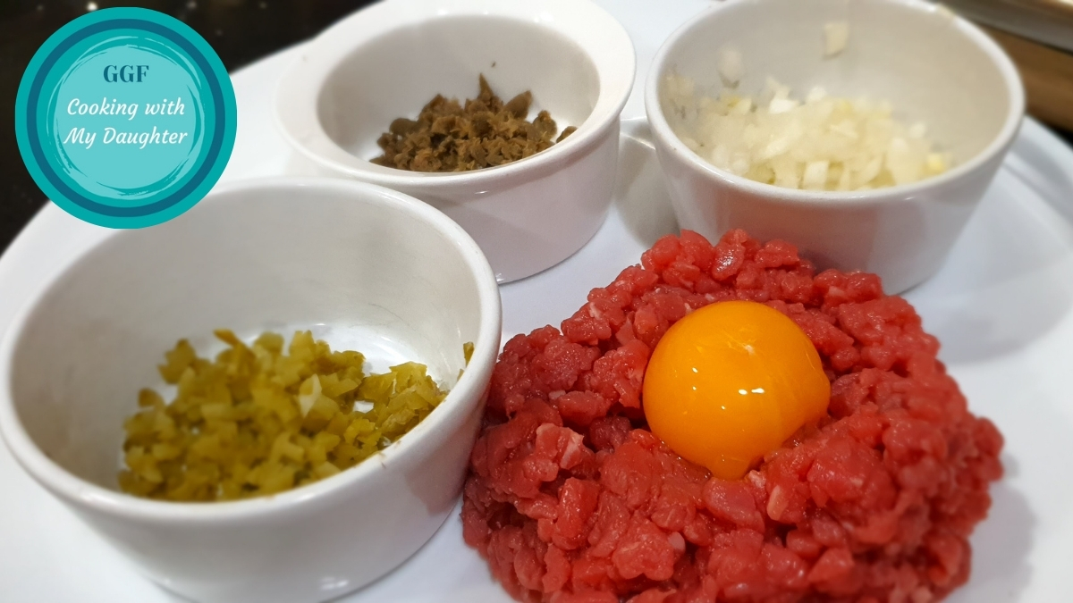 Tartare de boeuf au couteau. Image showing finely hand-chopped fillet steak with an egg yolk in the centre and small bowls containing the seasonings on the side - one with finely chopped gherkins, one with finely chopped capers and the third with finely chopped onions.